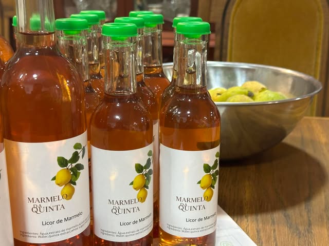 Garrafas de licor de marmelo com tampa verde e rótulo Marmelo da Quinta; ao fundo, uma taça com marmelos.